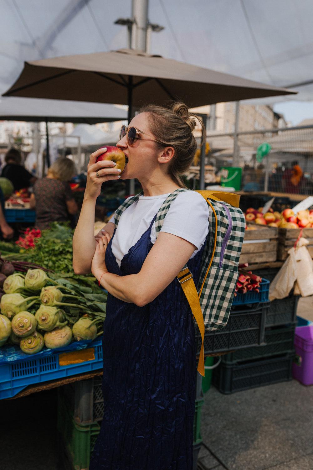 young woman eating apple while shopping with green gingham backpack. checkered laptop backpack by bettys home