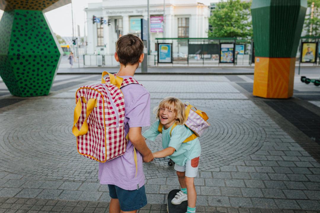 Two brothers laughing while skateboarding with gingham backpacks by bettys home. checkered school backpack for kindergarten