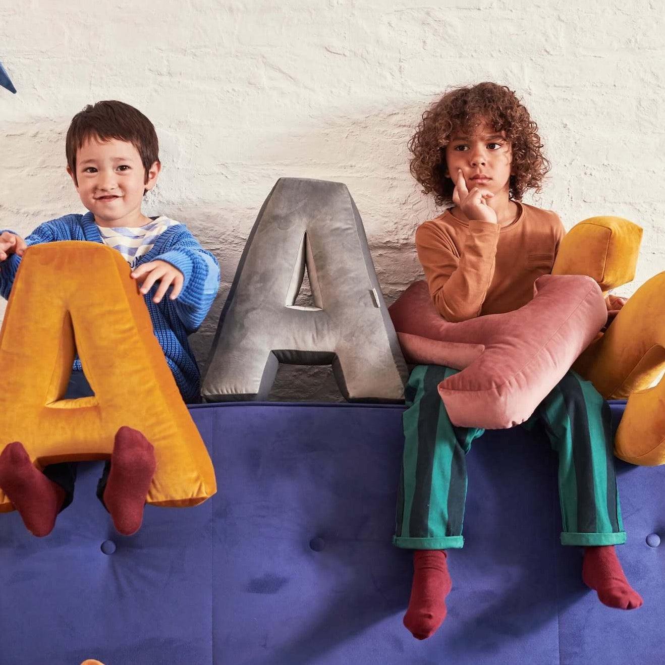 two boys sitting on couch with Velvet letter cushions by Bettys Home in hands