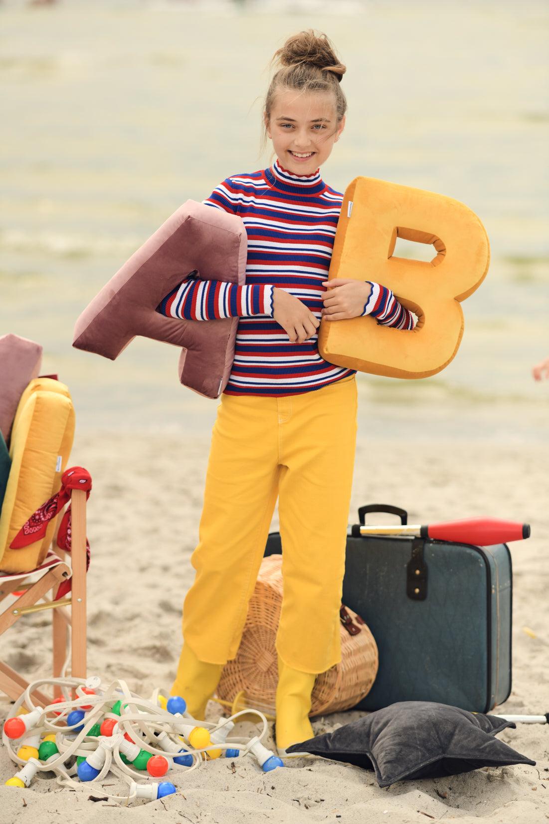 A cute teenage girl on the beach holding a velvet letter cushion B in yellow and A in pink. Baby shower decorations for a boy.