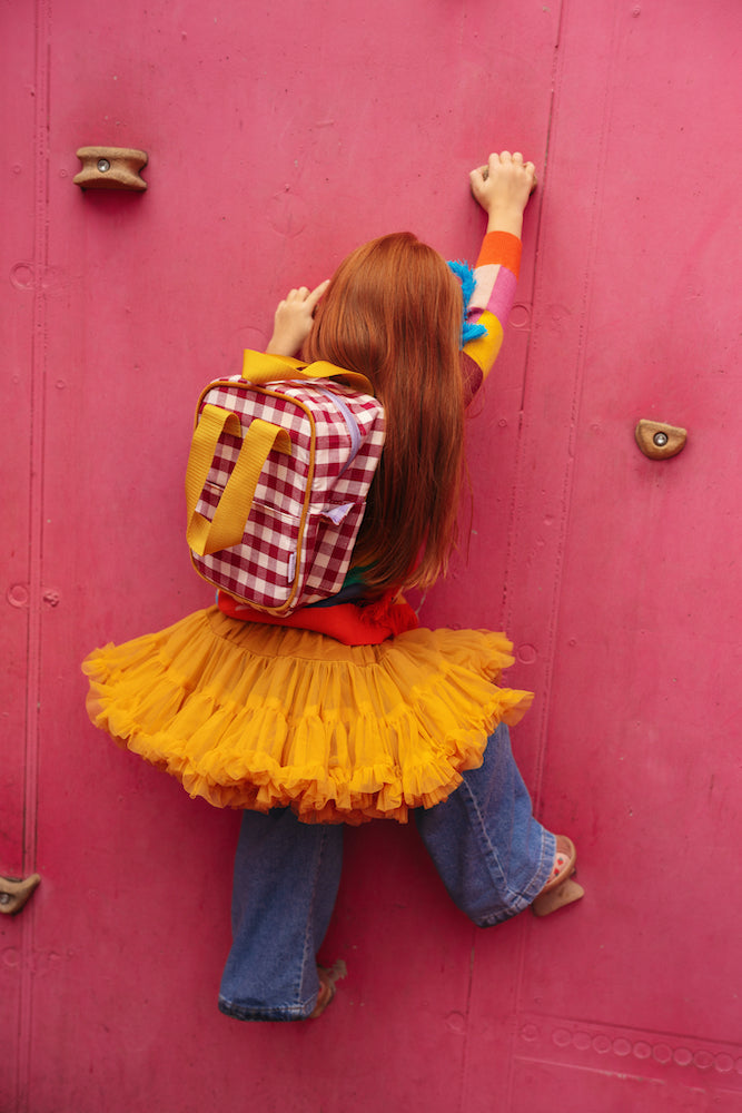 A little girl climbing a pink wall in a playground with a gingham backpack by bettys home 