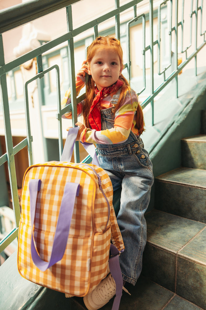 little girl standing on stairs at school with gingham backpack yellow by bettys home in hands. 