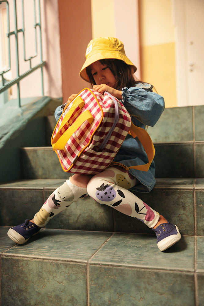 A little girl sitting on the steps at school with a gingham backpack in red by bettys home