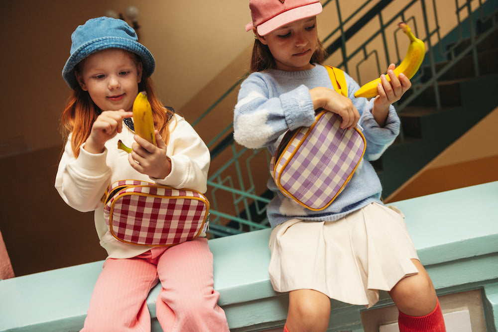 girls sitting in wall with banana and gingham fanny packs by bettys home. 