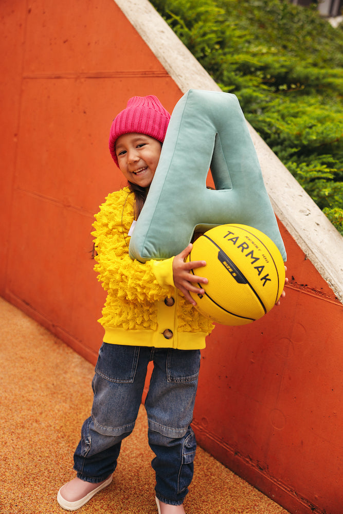 girl in the playground wearing a yellow jacket holds a yellow ball in her hands and a velvet letter pillow on it. letter pillow