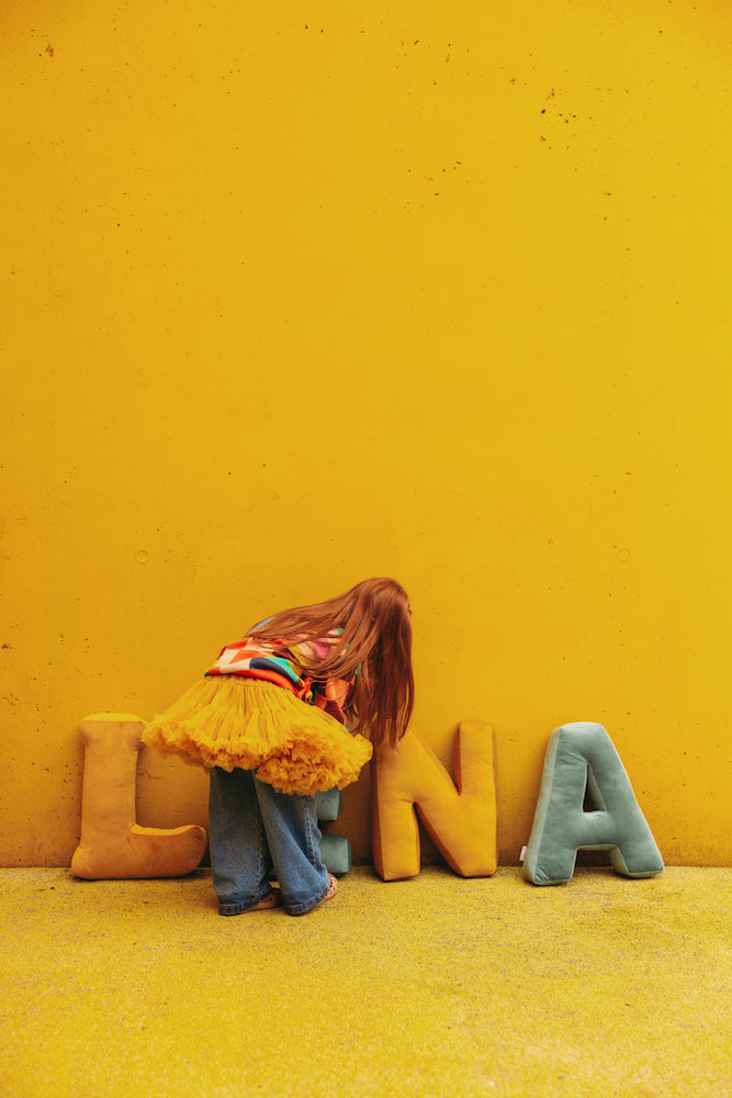 girl in playground against yellow wall stands next to velvet letter cushions LENA by bettys home.